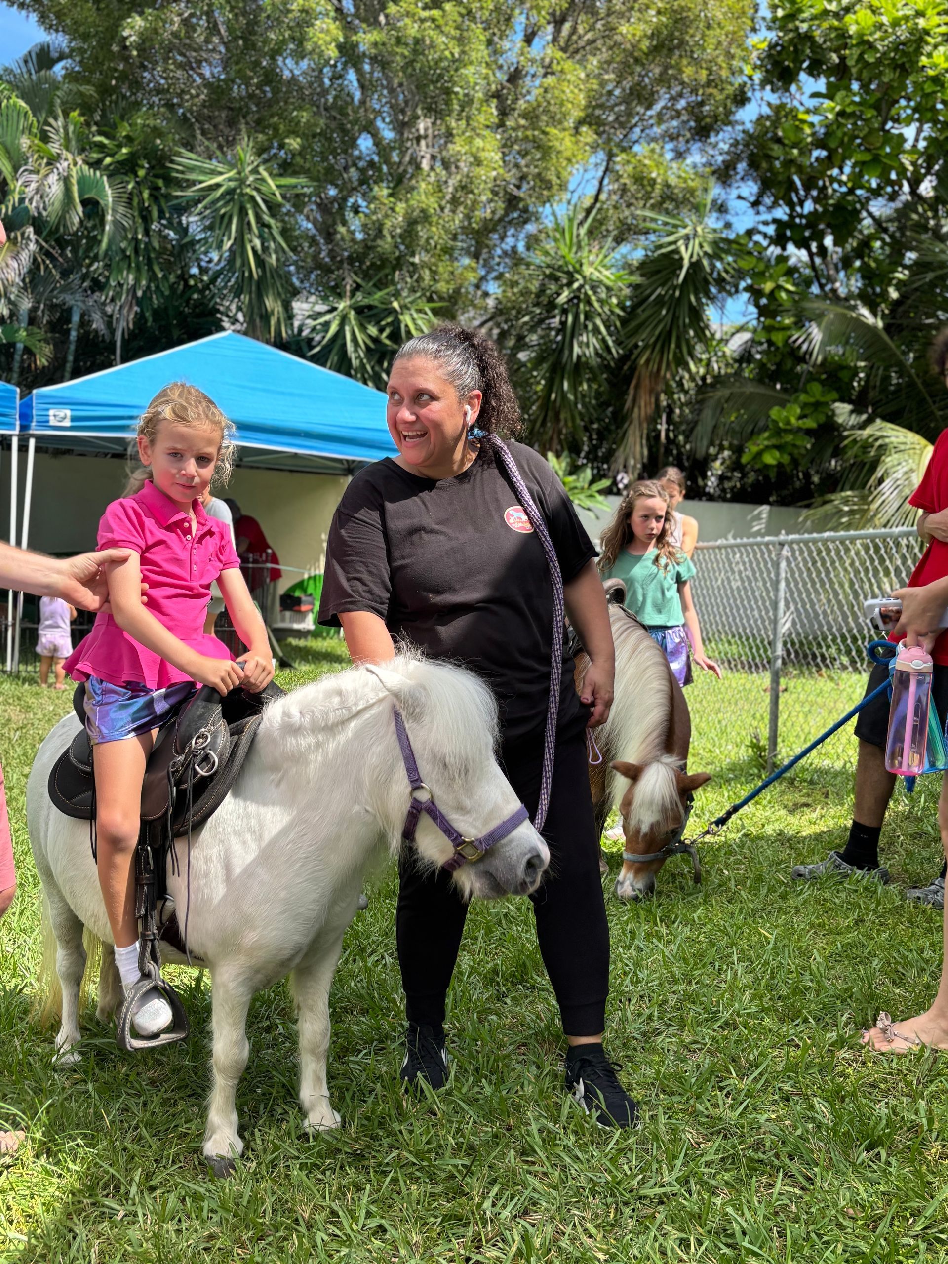 A child rides a white pony, guided by a woman. Others watch in a grassy area with tents.