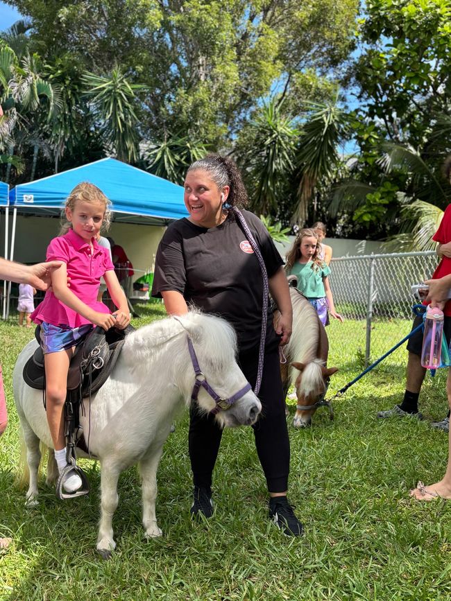 A child rides a white pony, guided by a woman. Others watch in a grassy area with tents.