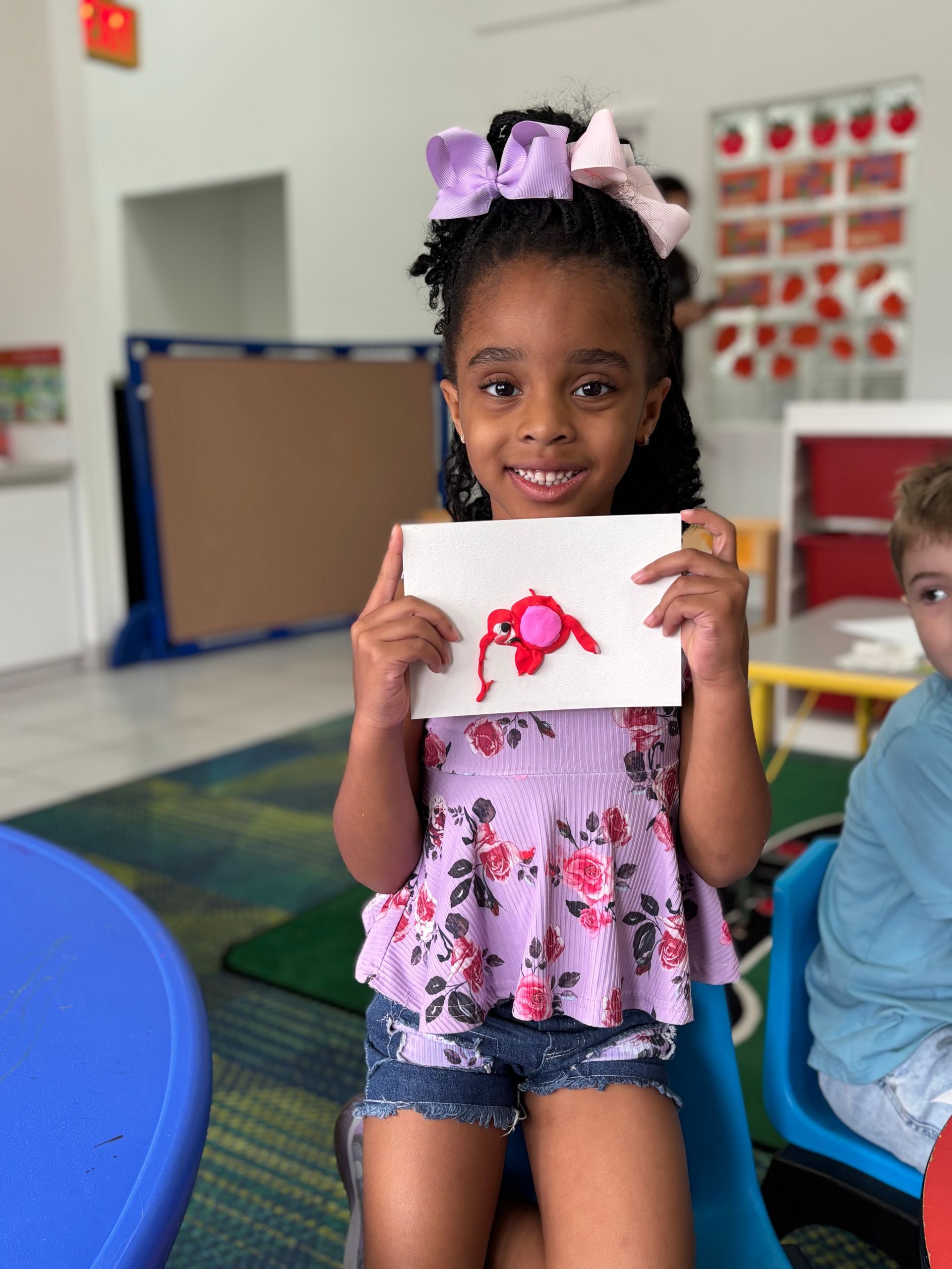 Young child holding a red artwork in a classroom, smiling.