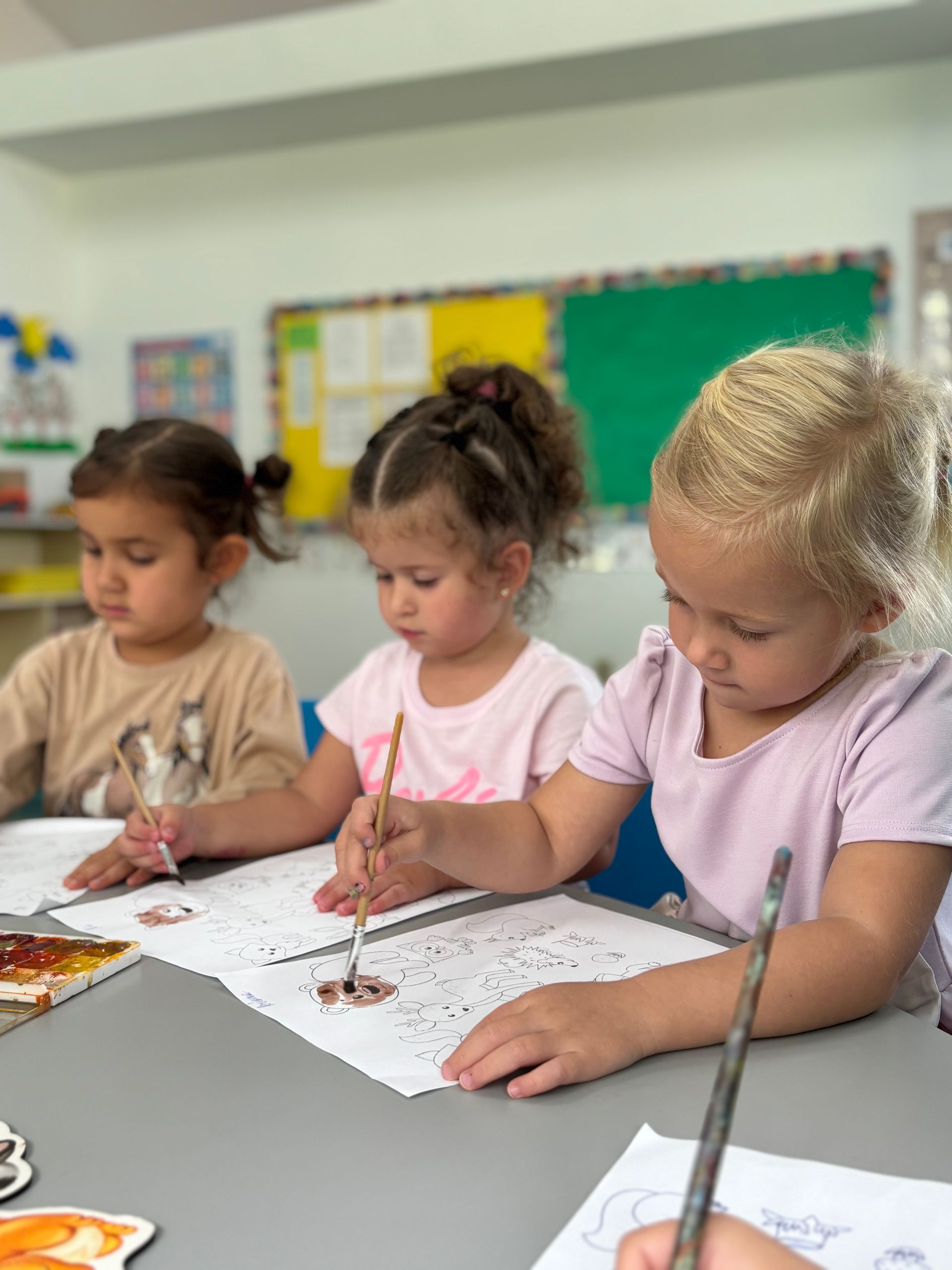 Three young children painting at a table, focused and concentrated, inside a brightly lit room.