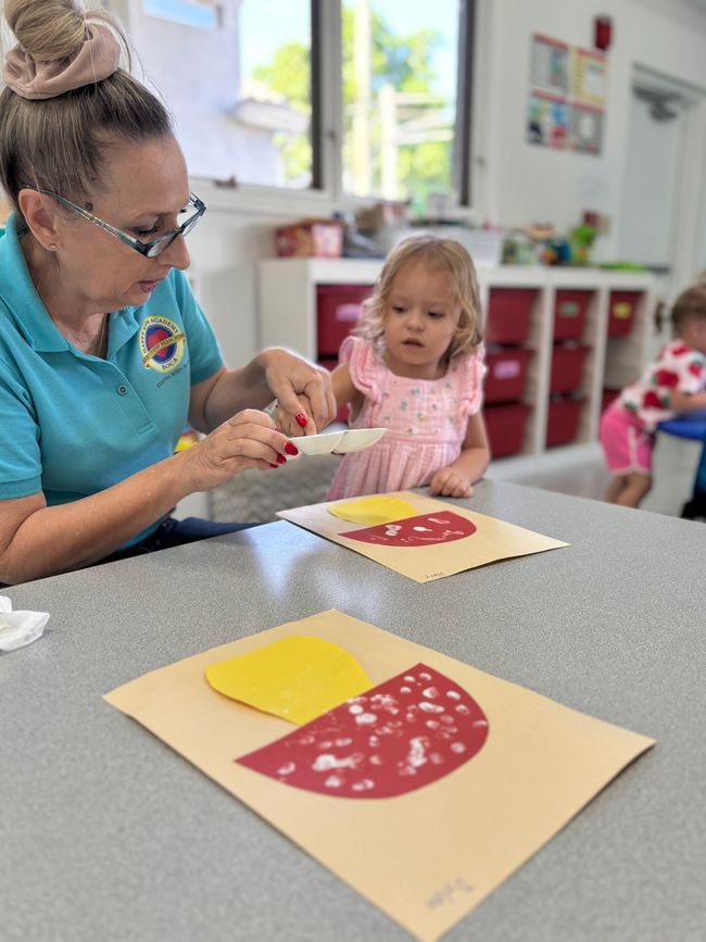 Teacher helping a child with a craft. They are making food shapes with paper.