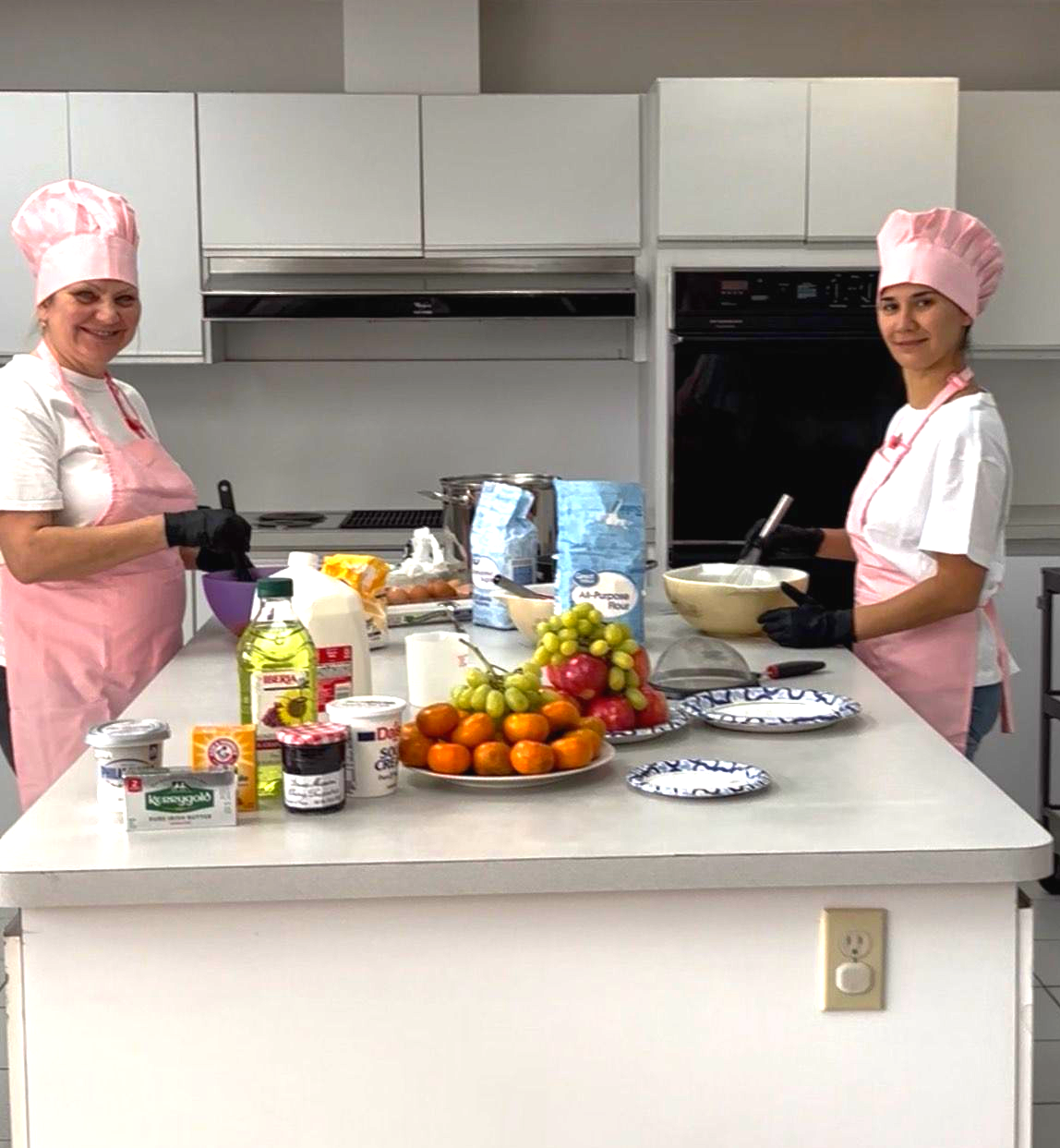 Two people in pink aprons and hats preparing food in a kitchen. Ingredients and fruit on a countertop.