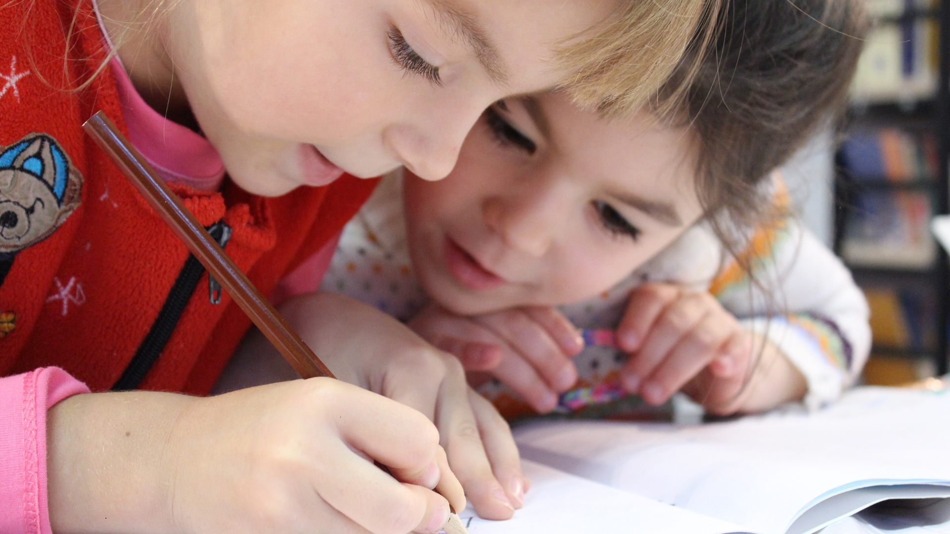 Two young children intently focused on a notebook, one writing with a pencil.