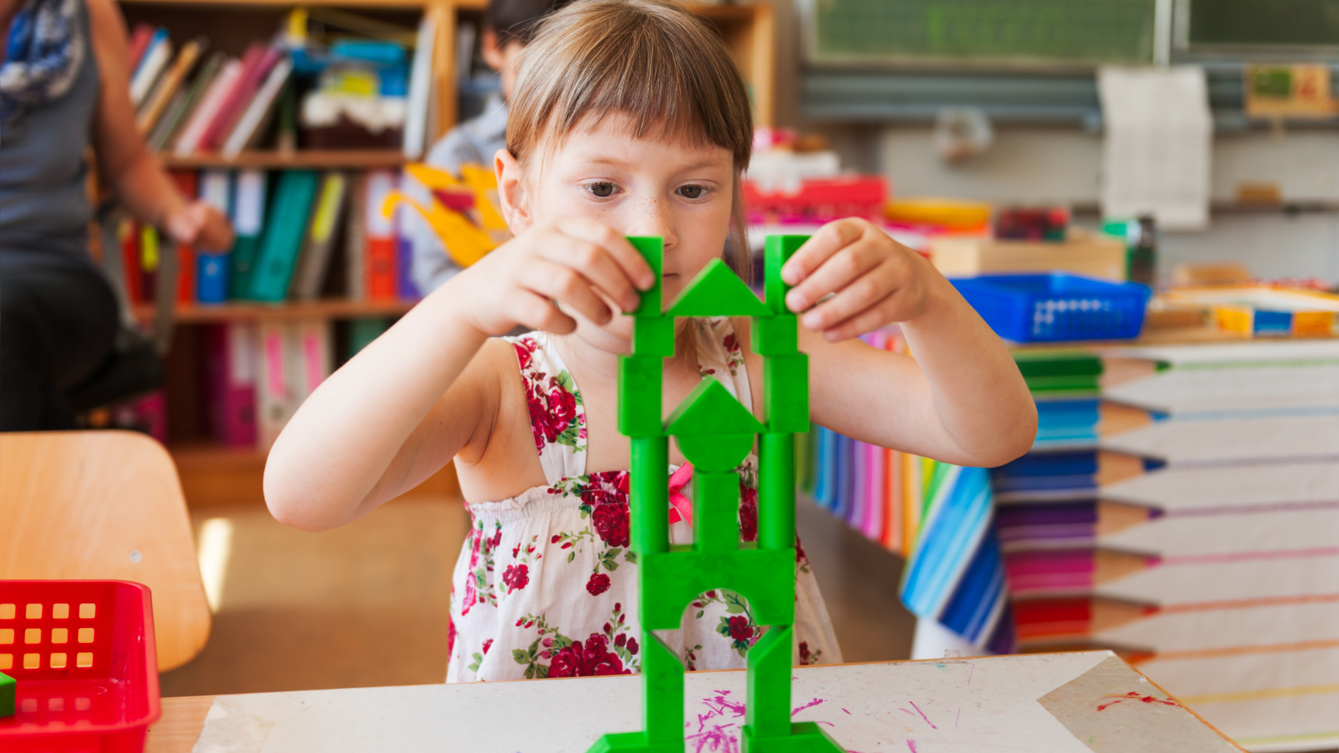Girl building a tower of green blocks at a desk in a classroom.