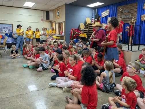 Children in red shirts sitting on the floor, clapping, with adults on a stage.