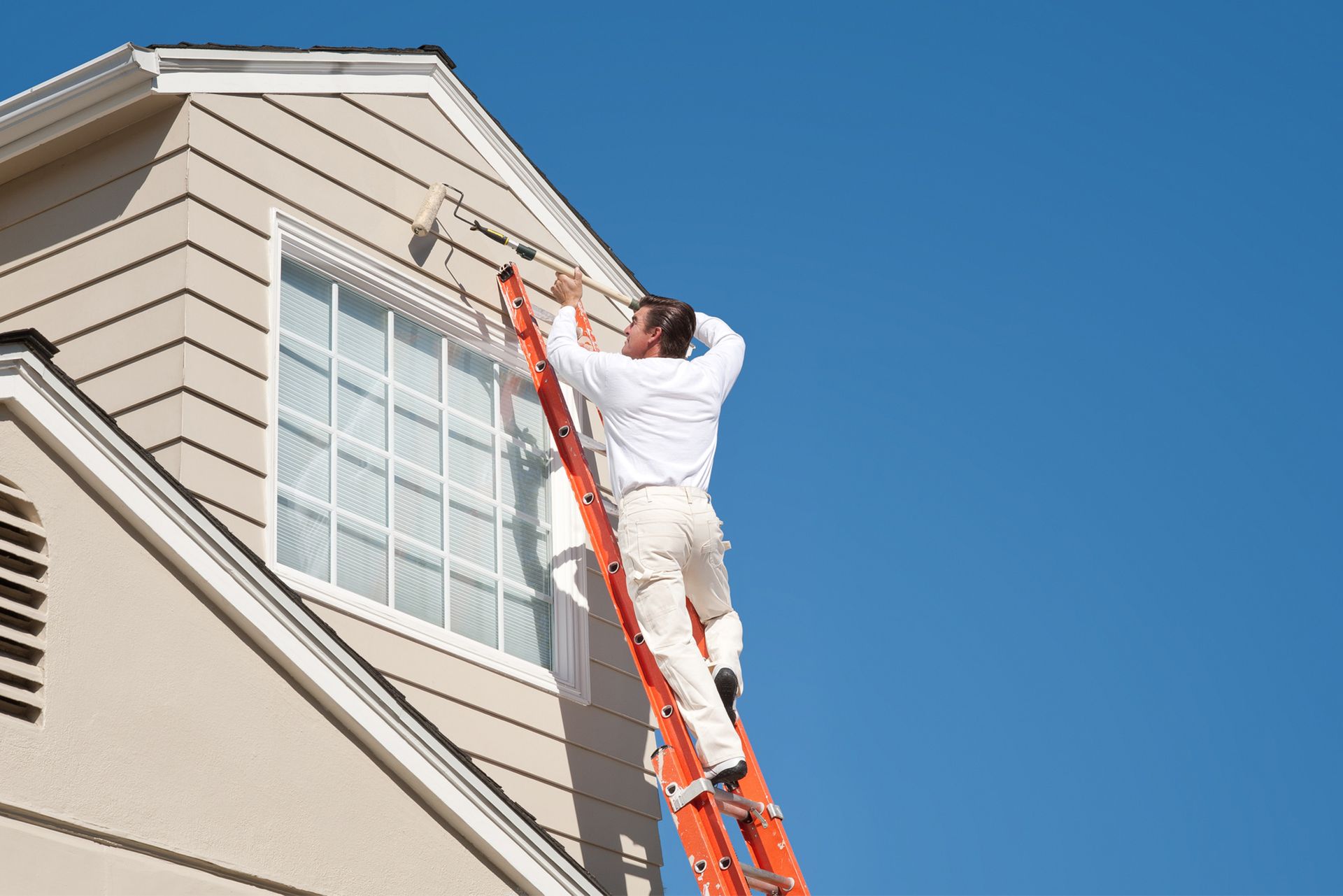 A man is standing on a ladder painting the side of a house.