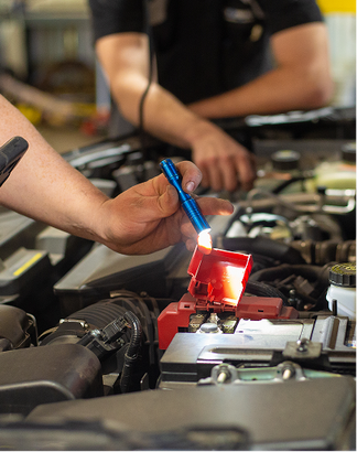 A smiling mechanic wearing a cap and hoodie works under the open hood of a car in a workshop. | Performance Automotive Repair