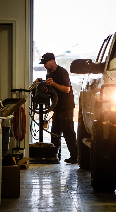 A person in dark clothing works on a piece of equipment in a garage next to a large vehicle. | Performance Automotive Repair