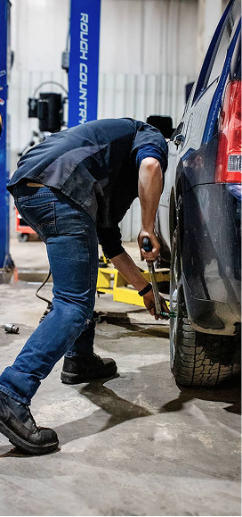 A mechanic wearing dark clothing works on the tire of a vehicle inside a garage with a lift in the background. | Performance Automotive Repair