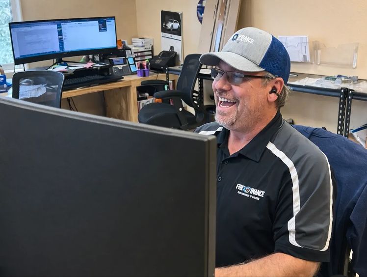 A smiling person wearing a cap and collared shirt works at a desk with dual monitors in an office setting. | Performance Automotive Repair