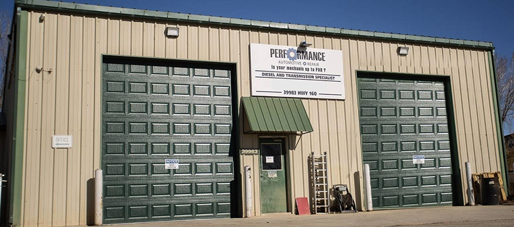 A beige metal commercial building with two large, dark green garage doors and a central entrance under a small awning. | Performance Automotive Repair