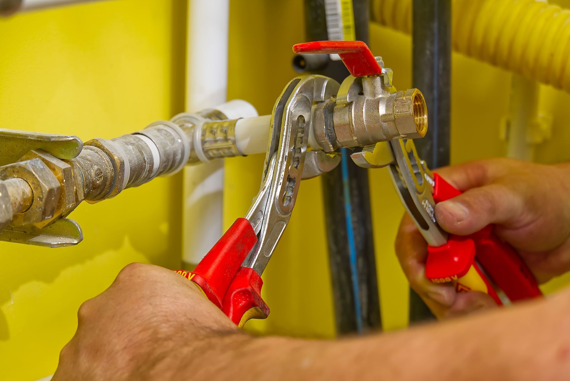Hands using red-handled pliers to tighten a pipe fitting in a yellow utility area