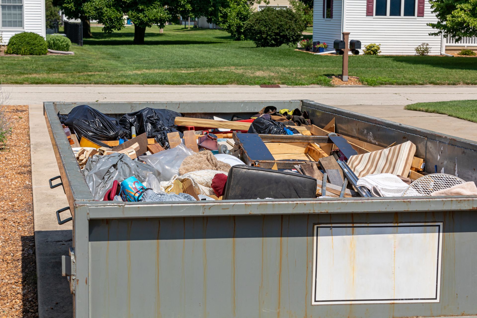 A large metal dumpster filled with discarded household items sits on a concrete surface in front of a grassy lawn and residential neighborhood.
