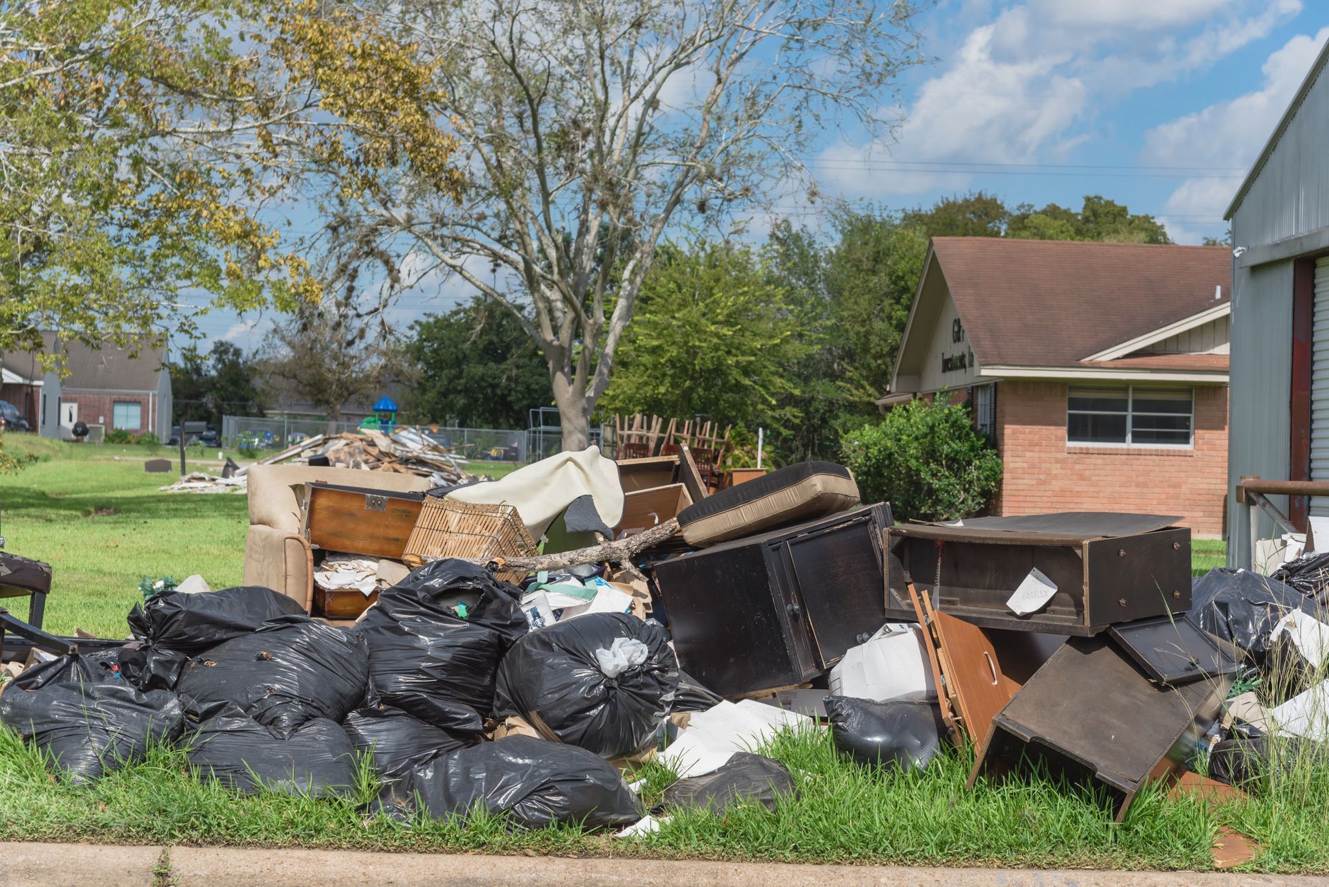 Pile of trash bags, furniture, and debris on a grassy lawn in front of houses. Sunny day with a tree in the background.