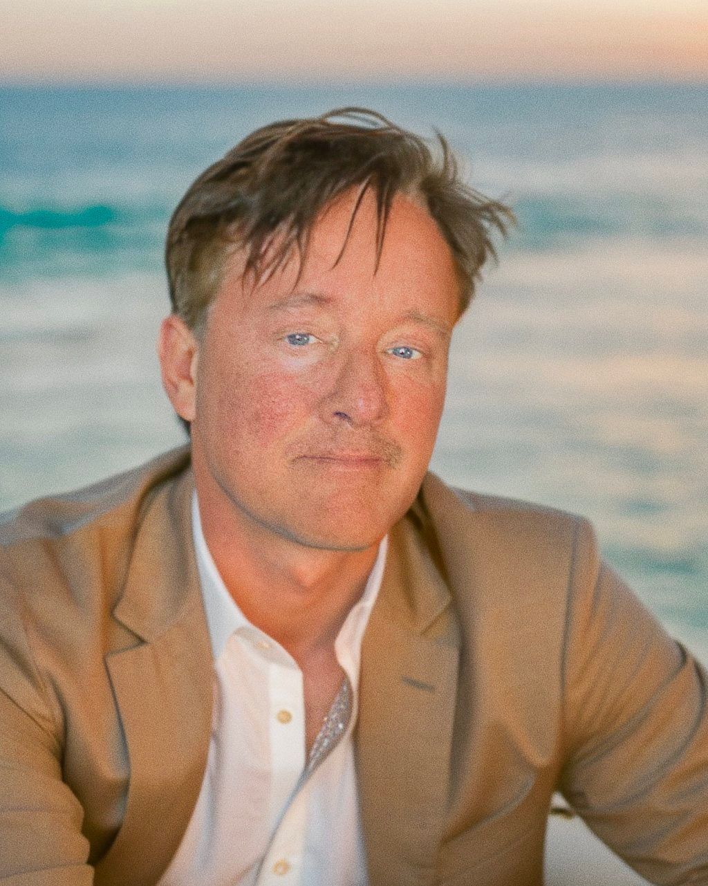 A man in a suit and white shirt is sitting in front of the ocean.