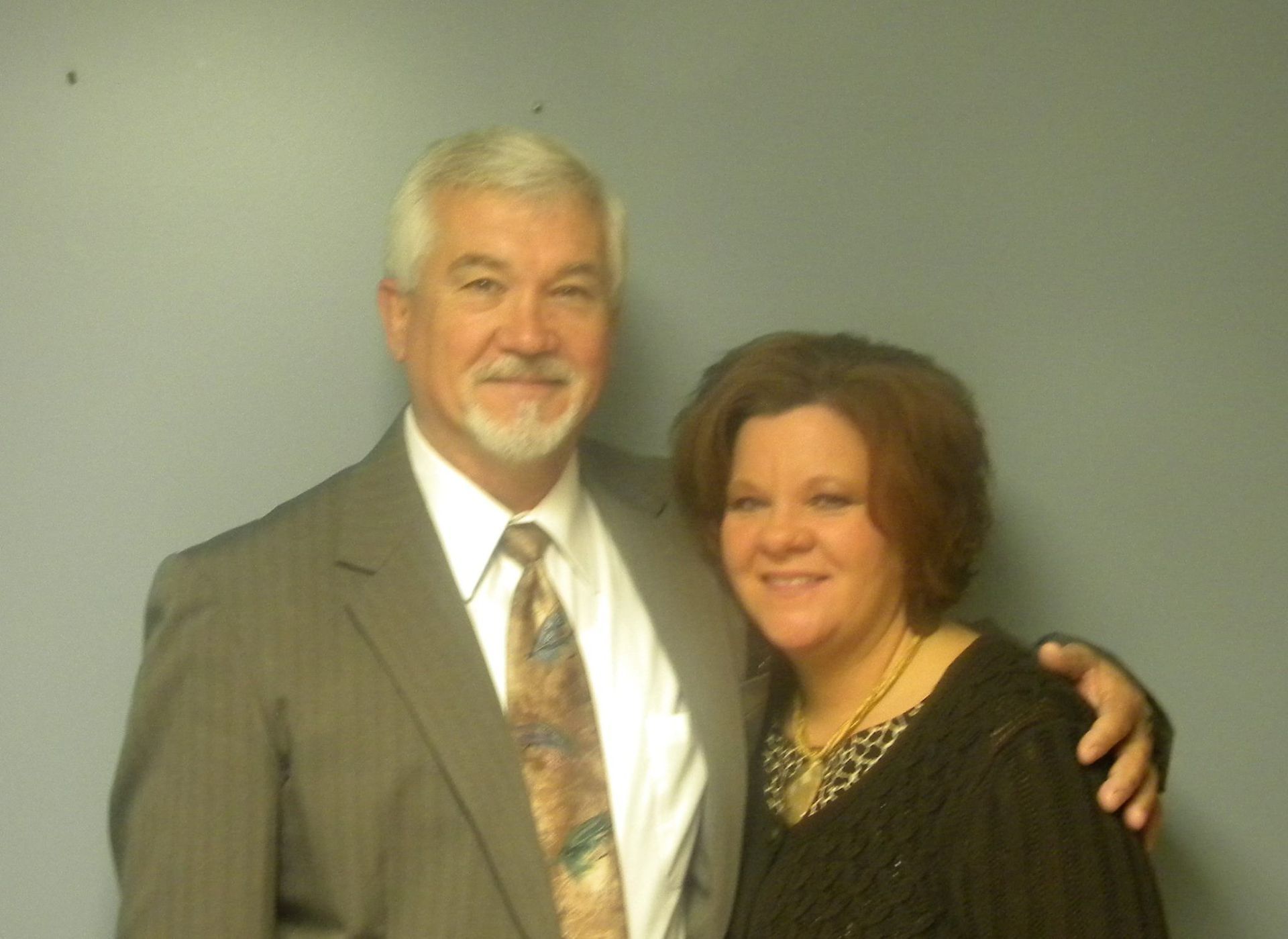 Man in suit and woman with arm around him, smiling in front of a blue wall.