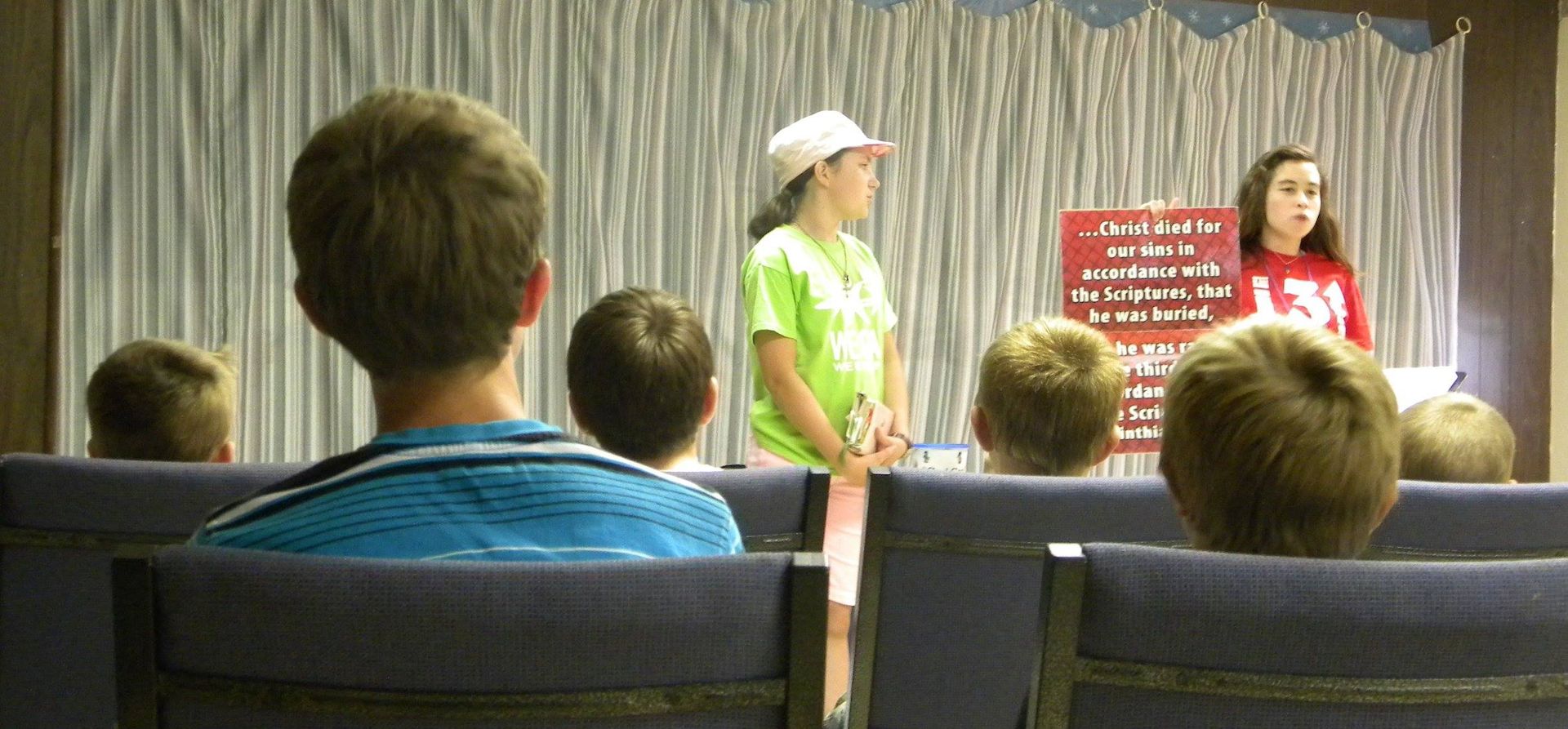 Children in a church auditorium, two girls at the front, one with a sign, the others seated.