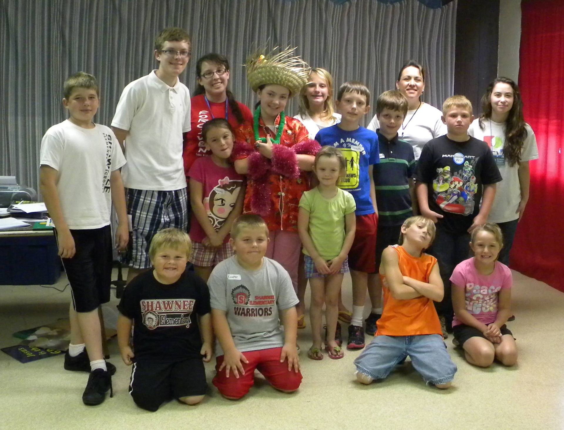 Group of kids smiling, posing for a photo. Some are wearing festive outfits. Indoors, against a backdrop.