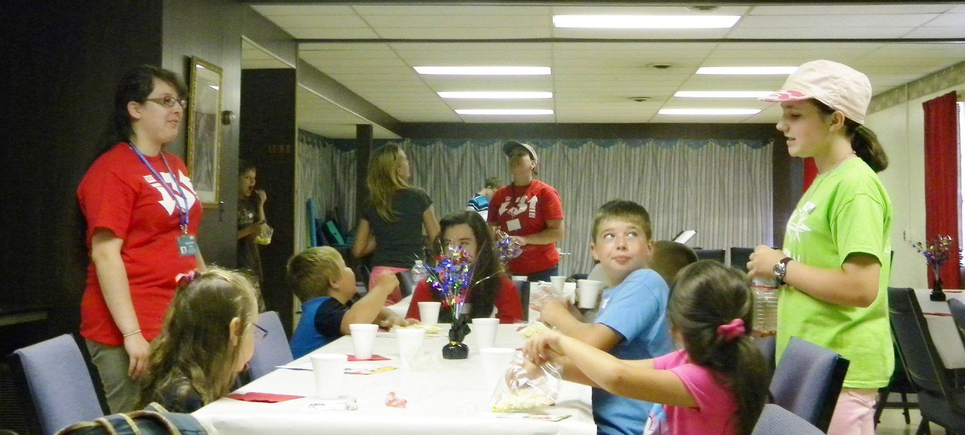 Children and adults at a party; indoors, a long table is set with cups, snacks. People wear red and green shirts.