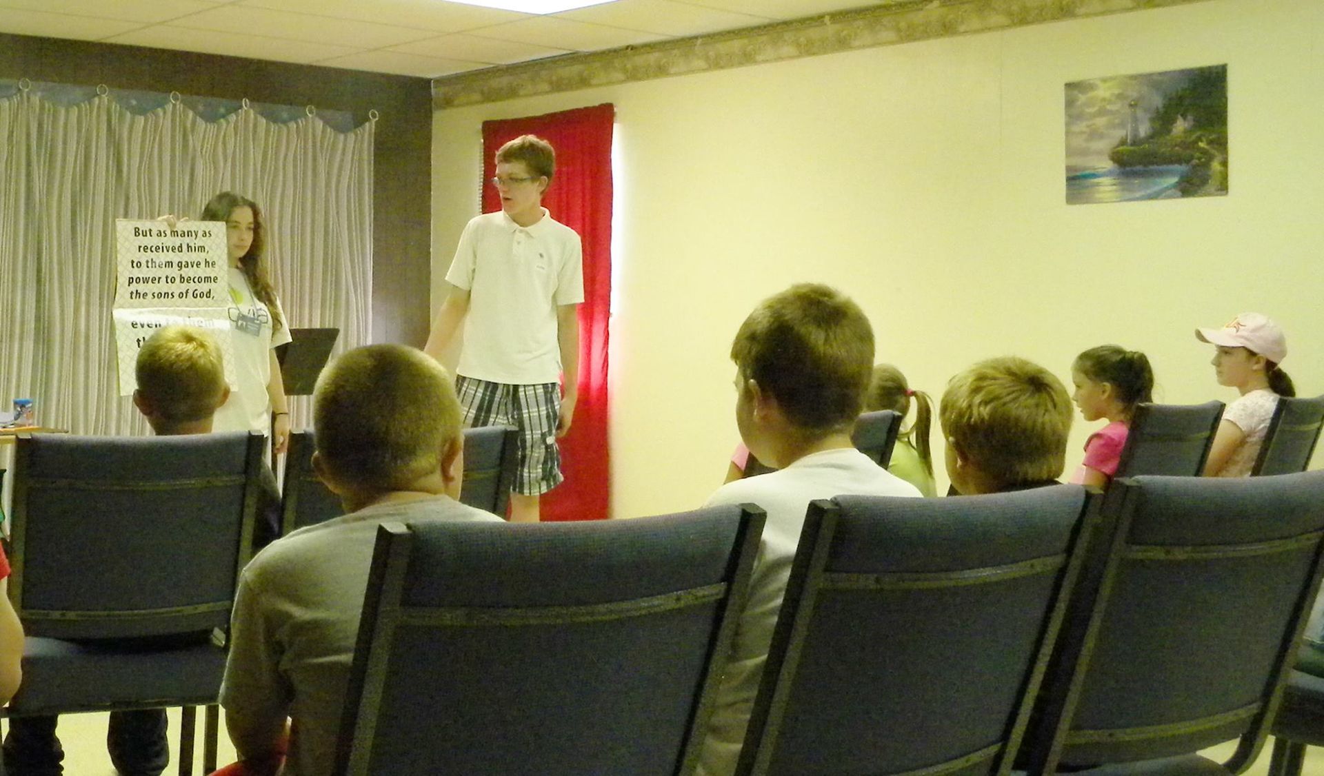 Children in a room watching a presentation; two youth stand near a poster, one is speaking.