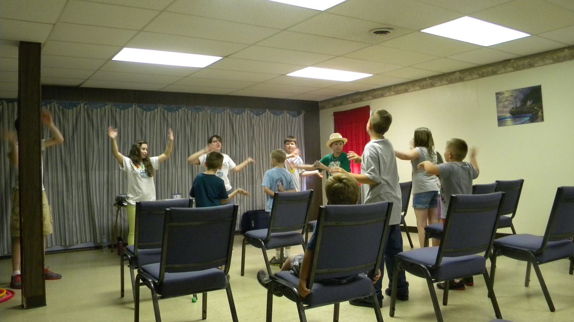 Children dancing in a room with chairs; some have hands raised.