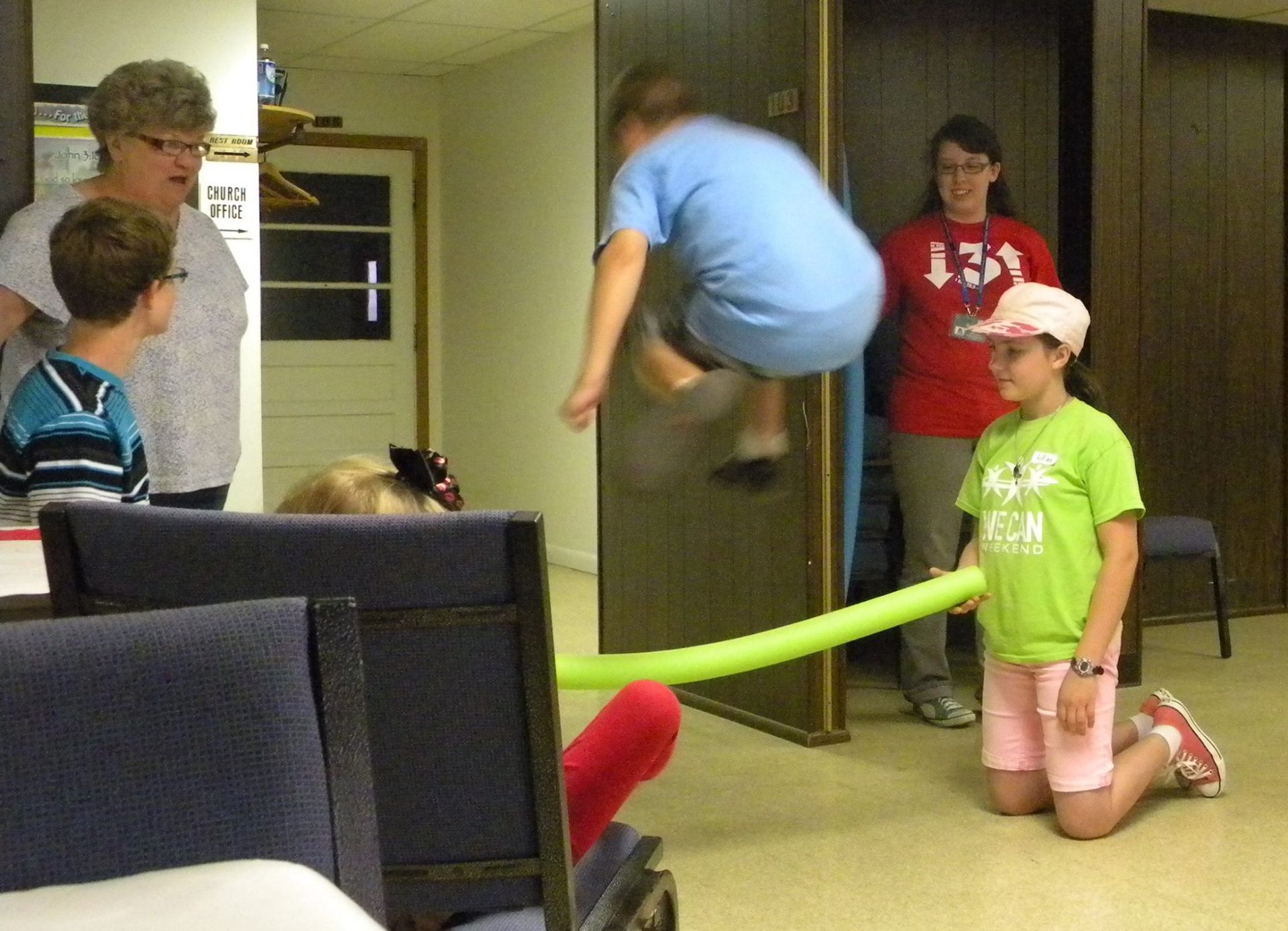 People playing a game: boy jumps over a balloon rope, others watch. Indoors, some wear casual clothes.