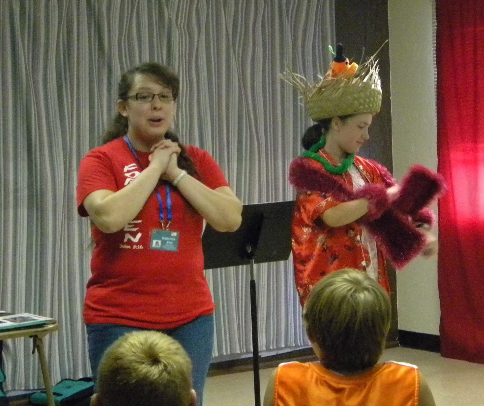 Two women on stage; one in red shirt, hands clasped, other in Hawaiian attire; audience in foreground.