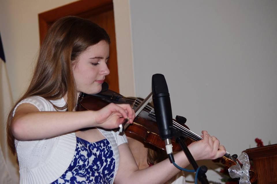 Woman playing a violin into a microphone, indoors, wearing a white sweater over a blue and white dress.