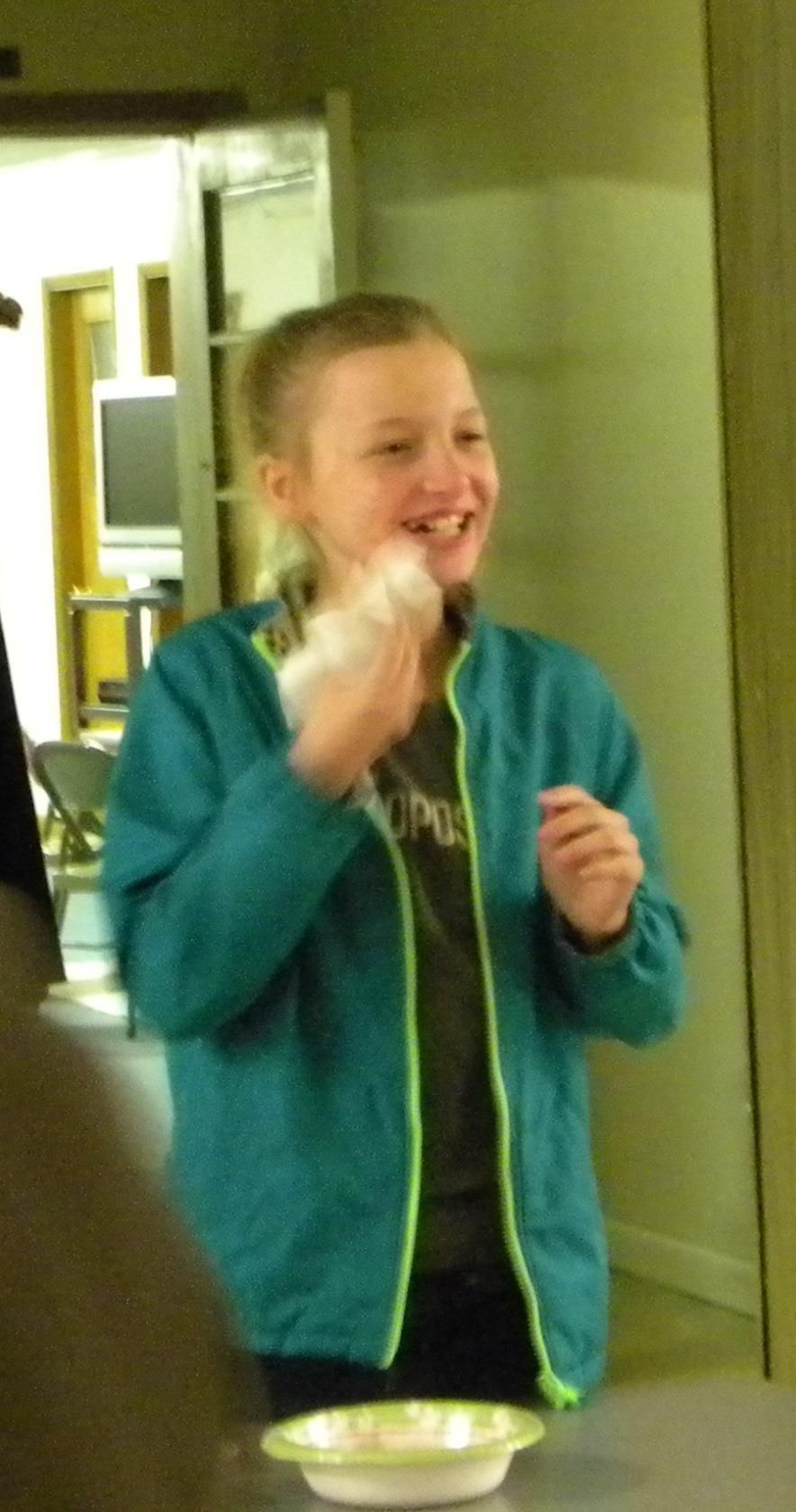Young person laughing, holding a white object over a pie plate; wearing a turquoise jacket in a room.