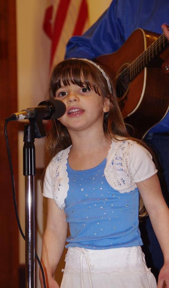Young girl singing into a microphone, wearing blue shirt and white shrug, guitarist in background.