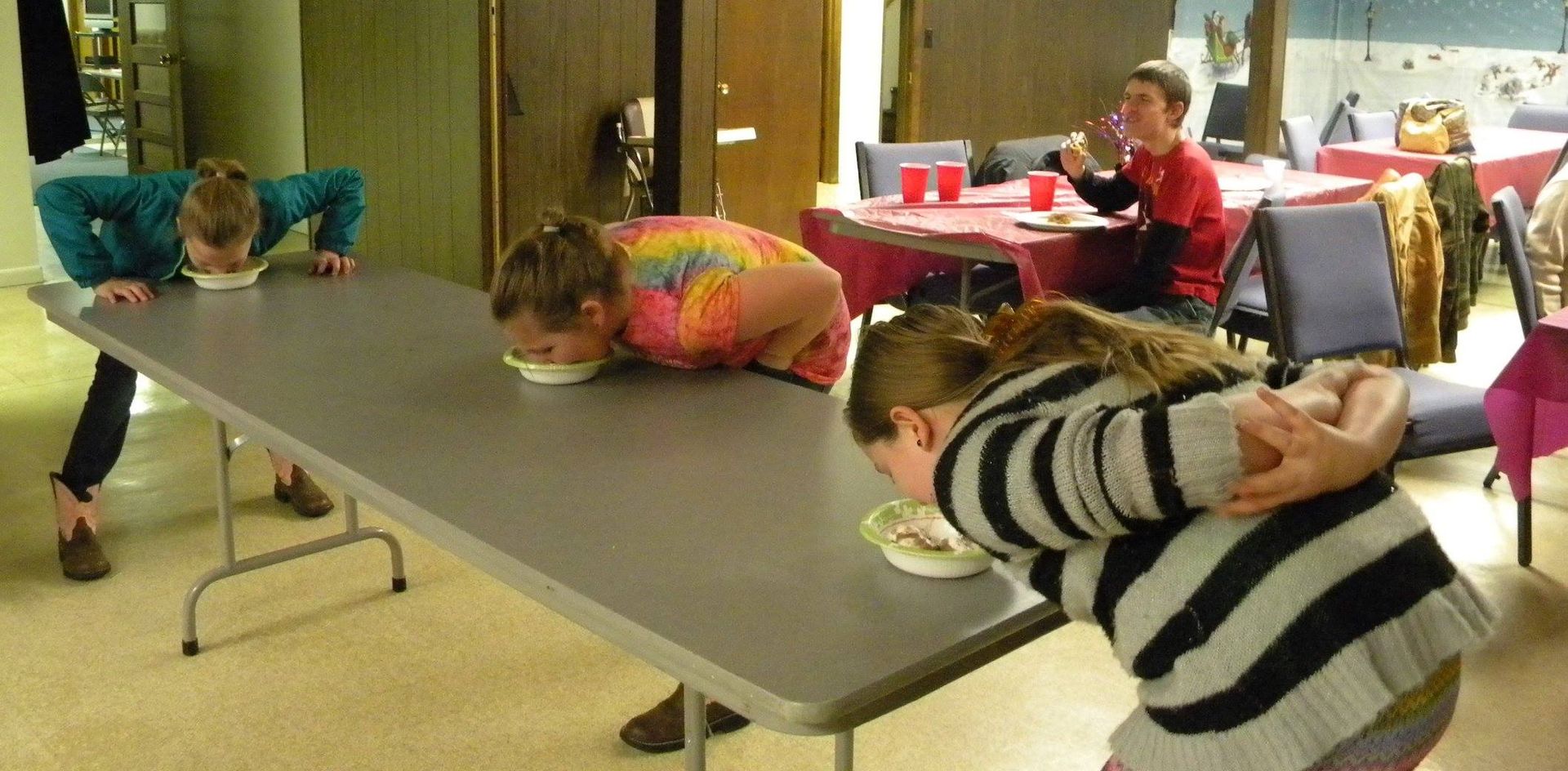 People eating from bowls on a table, with their bodies arched over the table. A person watches in the background.