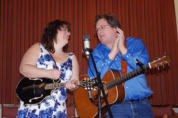 Woman plays mandolin; man plays guitar, singing at a microphone. Stage with brown curtains.
