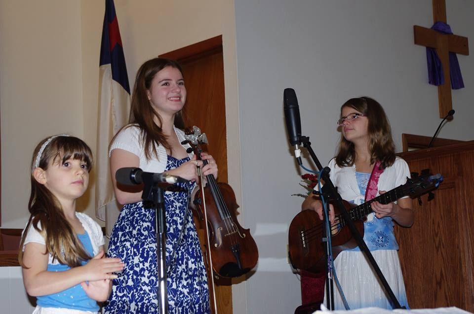 Three people with instruments perform in a church. A woman holds a violin, while a girl has a ukulele.