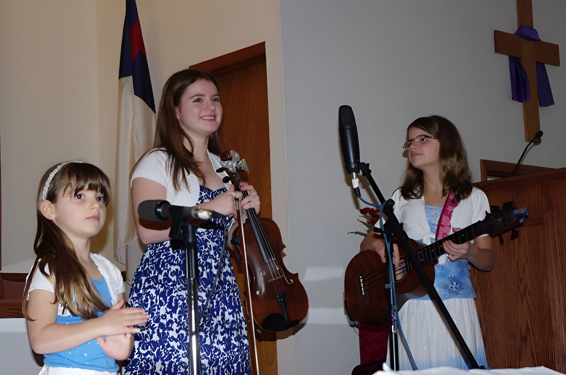 Three young musicians performing on a stage. One with a violin, another with a mandolin, and the third clapping.