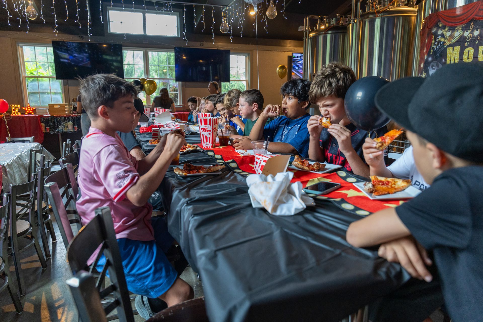 Children eating pizza at a birthday party, indoors. Black table cloth, balloons, and decorations.