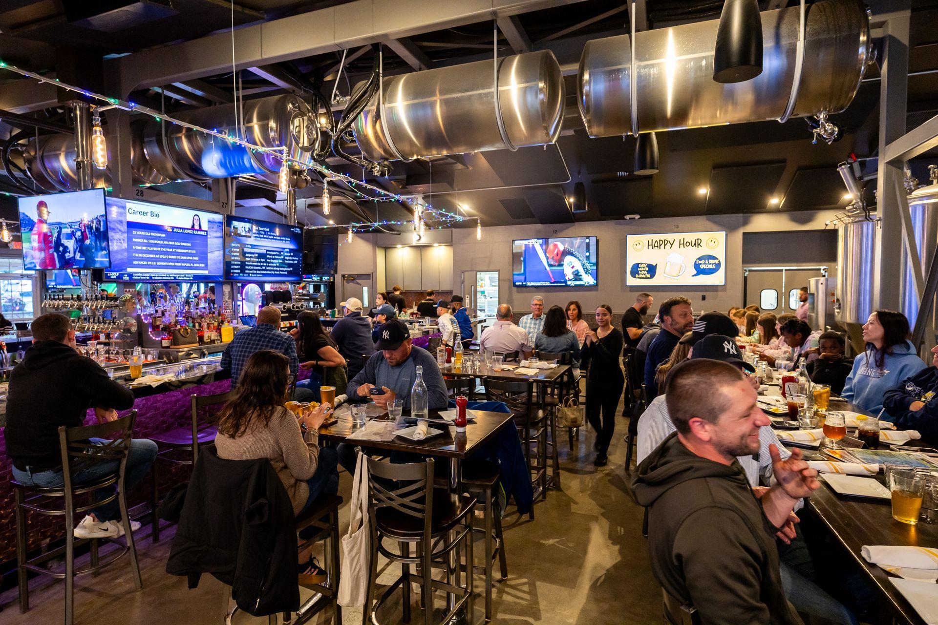 People at tables and bar in a busy modern bar, watching TVs. Metal decor, dim lighting.