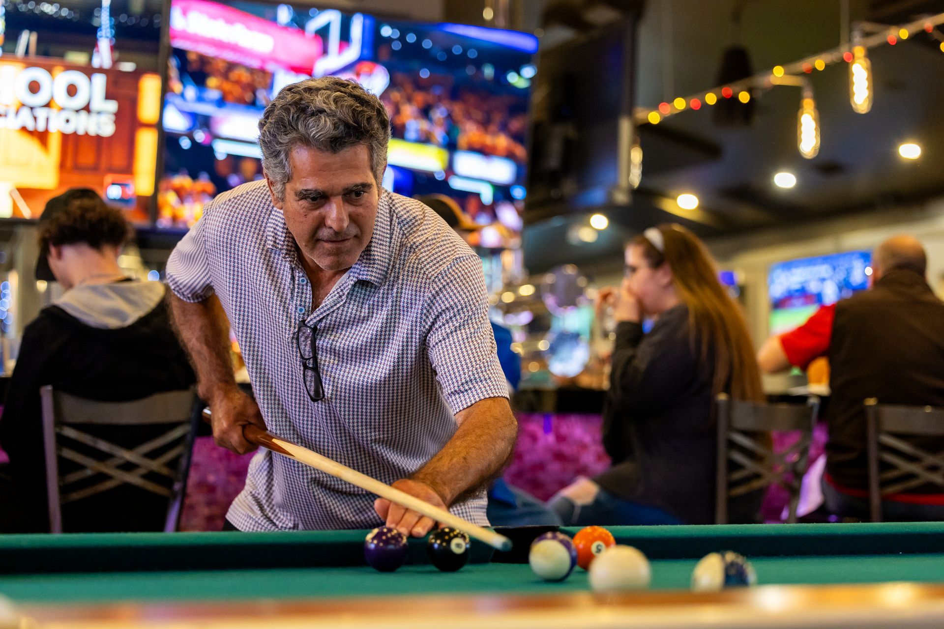 Man playing pool in a bar, aiming at the balls with a pool cue. Others watch and eat at the bar.