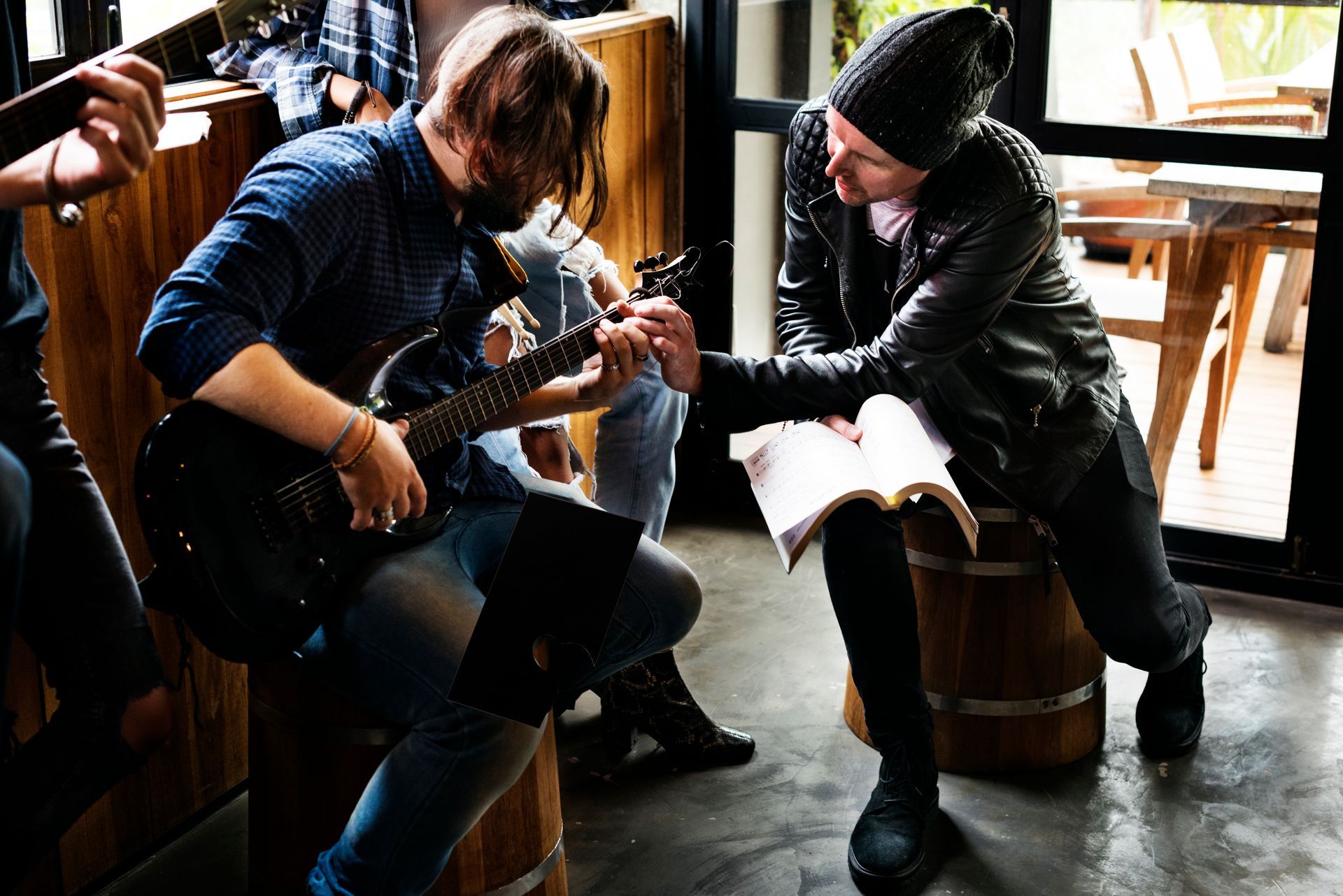 Two men in a room, one playing guitar, the other reading notes. Wooden barrel seats.