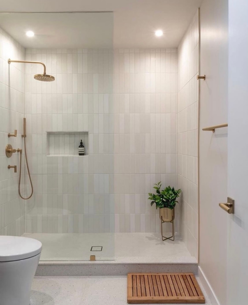 Modern bathroom with glass shower, gold fixtures, white tiles, and wooden mat.