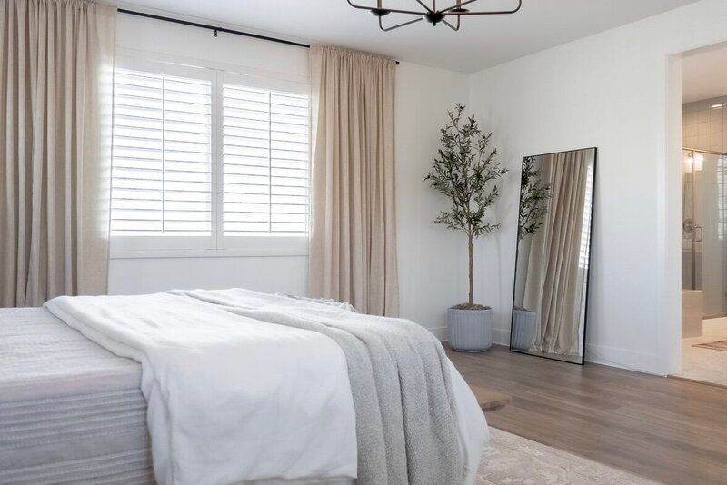 Bedroom with bed, tall mirror, shutters, curtains, and an olive tree. Neutral colors and natural light.