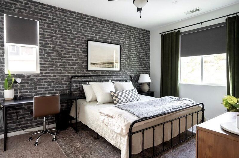Bedroom with brick accent wall, black bed frame, desk, and dark green curtains.