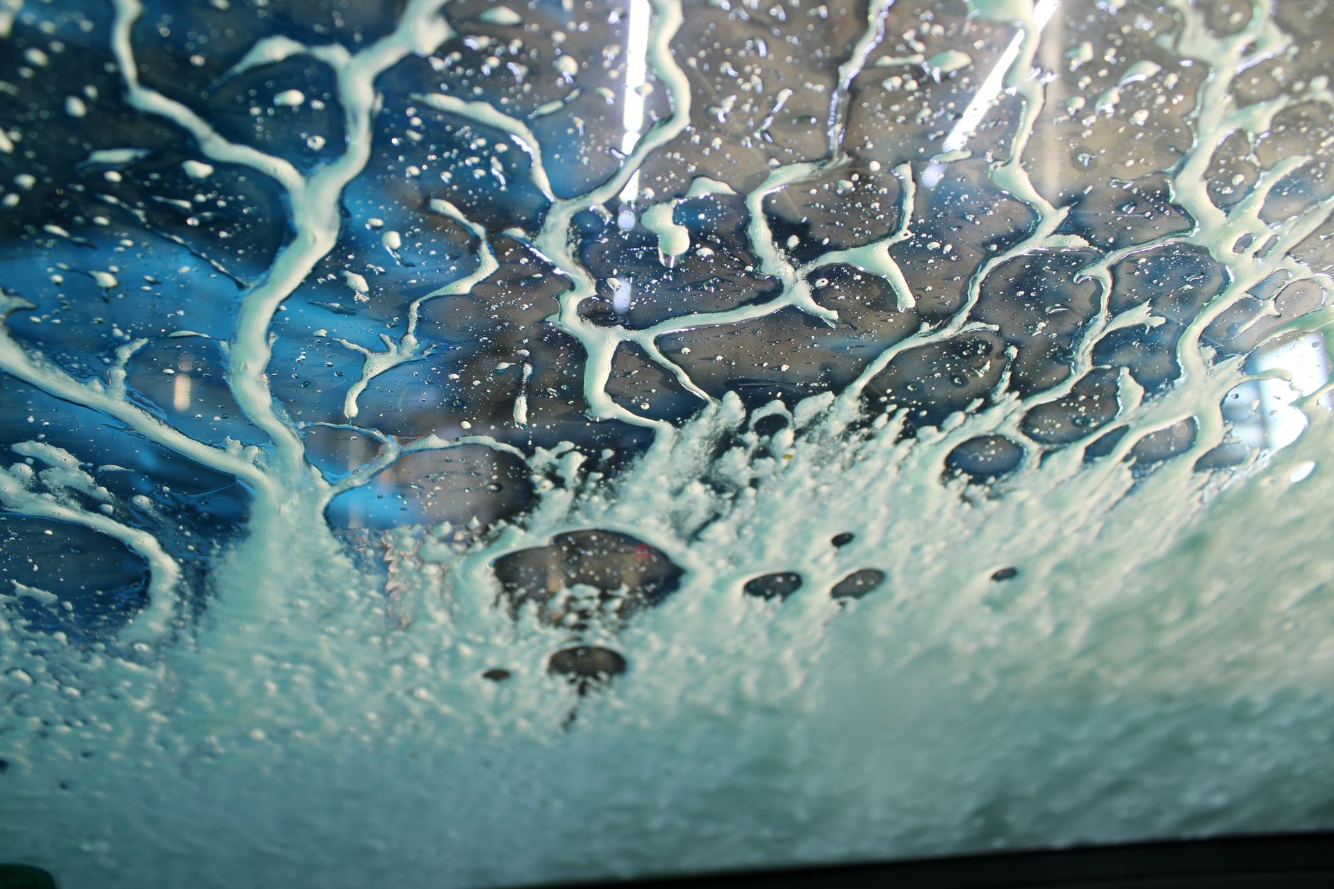 Inside a car wash, soapy foam coats the windshield, with a blue background visible through the wet glass.