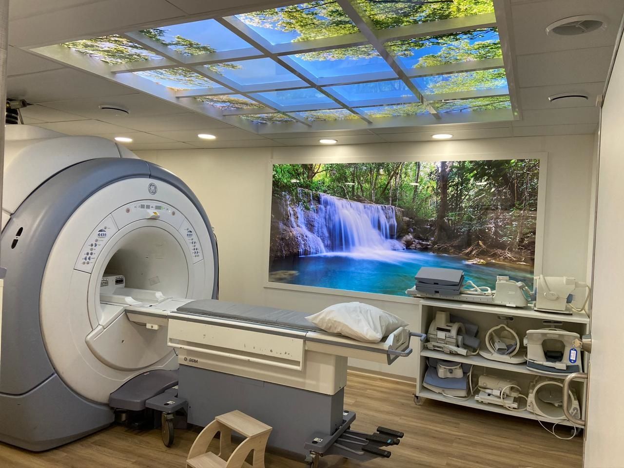 Aparelho de ressonância magnética em sala com painéis de parede e teto com temas da natureza; maca de exame presente.