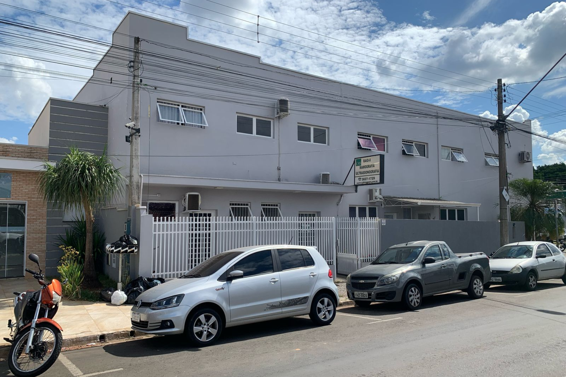 Prédio cinza com carros estacionados na frente, sob um céu nublado.