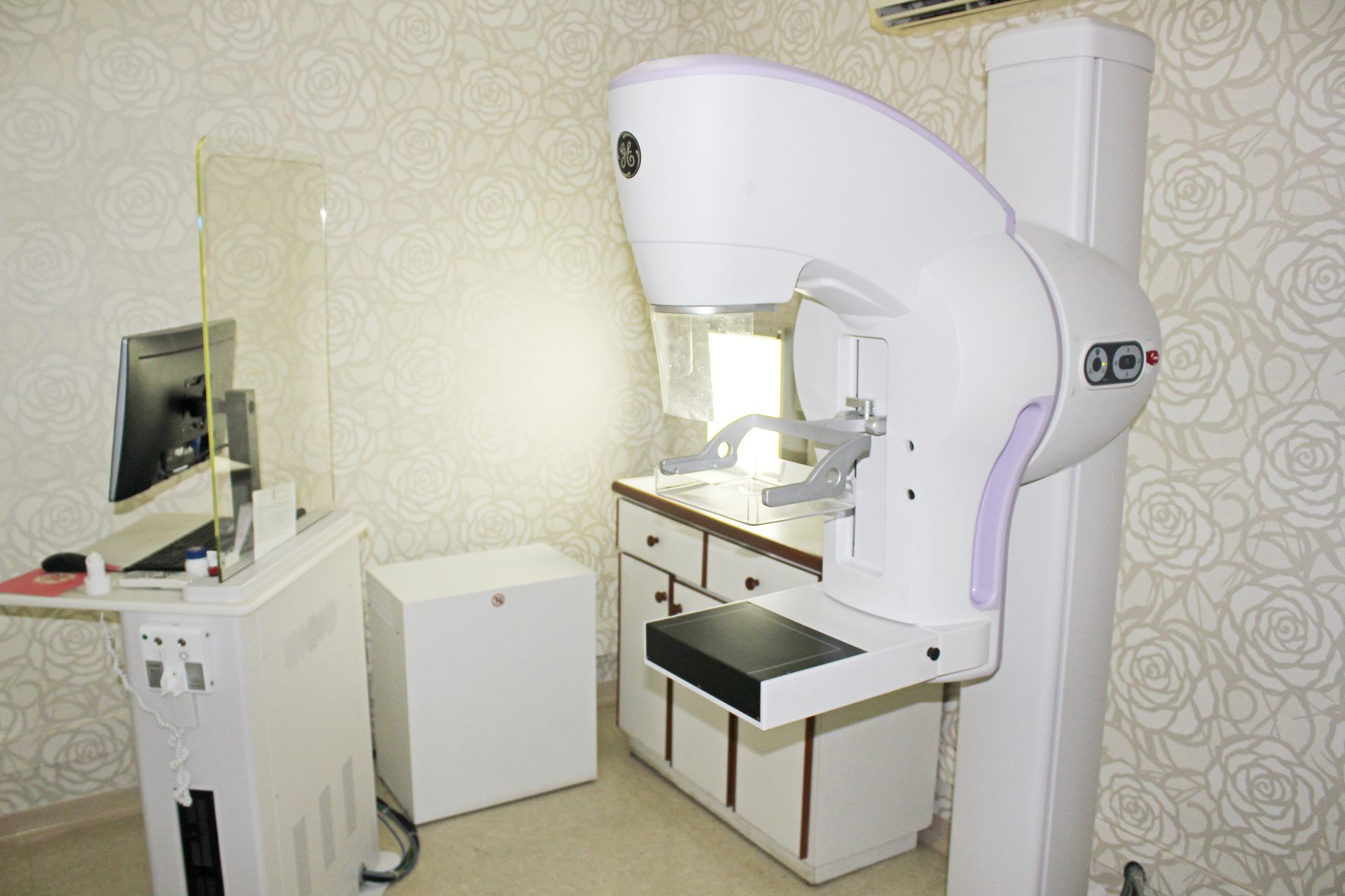 Aparelho de mamografia em ambiente médico. Também são visíveis um visor digital, um armário de armazenamento e uma proteção.