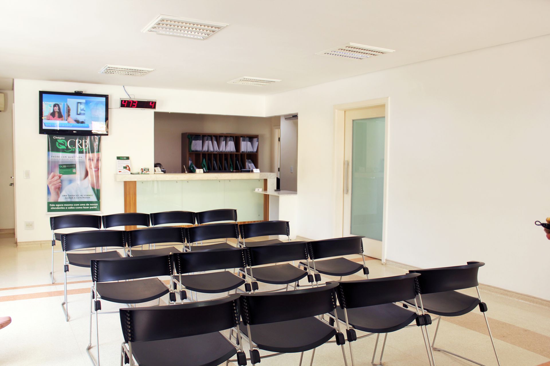 Sala de espera com cadeiras pretas em frente a um balcão de recepção; uma TV exibe um anúncio.