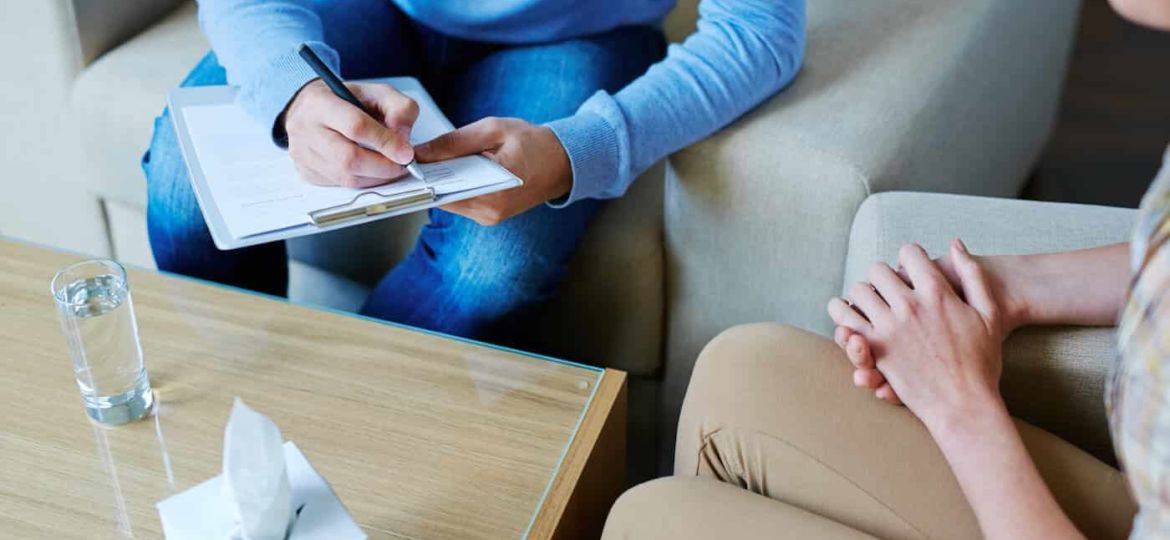 A man is sitting on a couch talking to a woman while holding a clipboard.