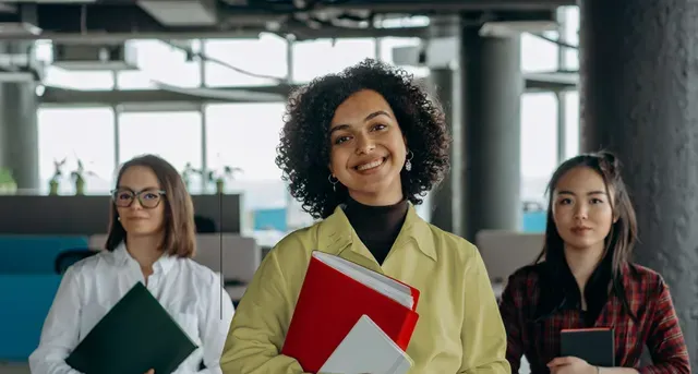 Three women are standing next to each other in an office holding books.