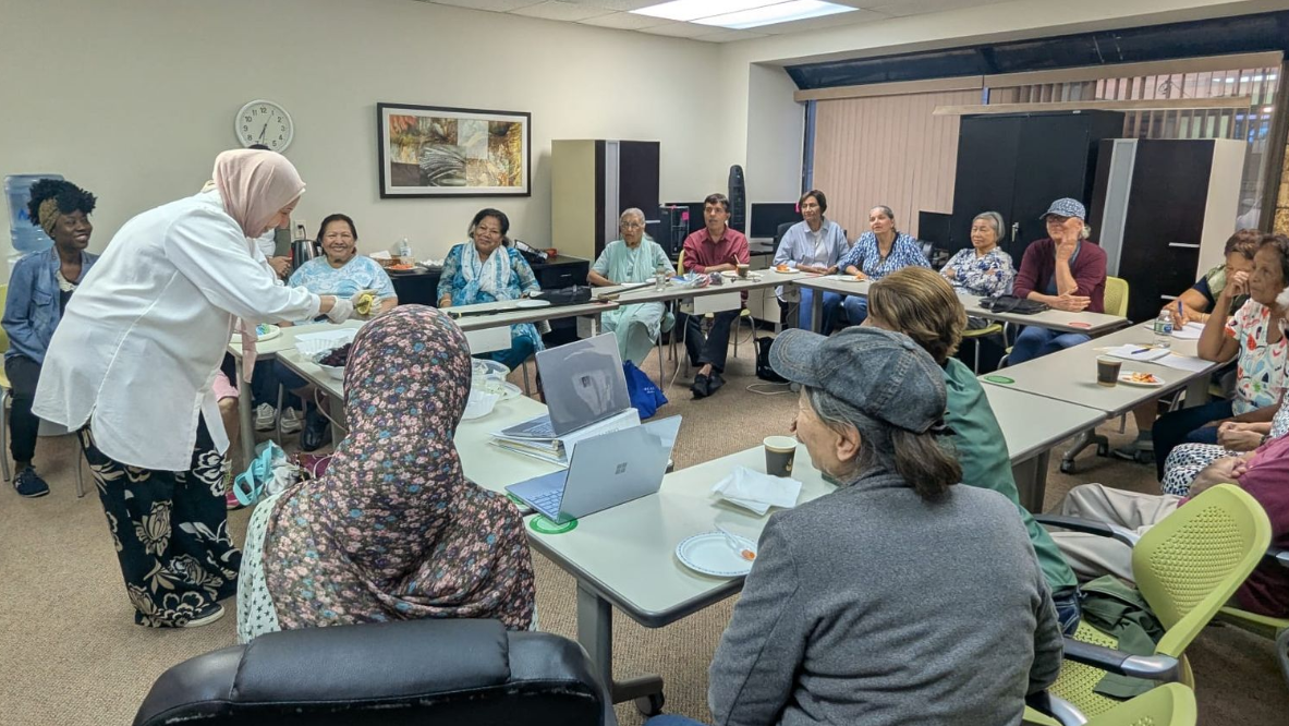Group of people in a meeting. A person in white is pouring liquid for seated individuals at tables.