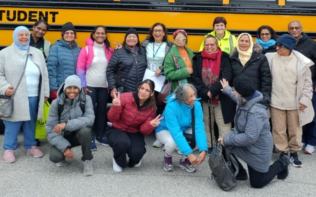Group of people posing in front of a yellow school bus. Most are wearing jackets, smiling, some crouched. Outdoors.